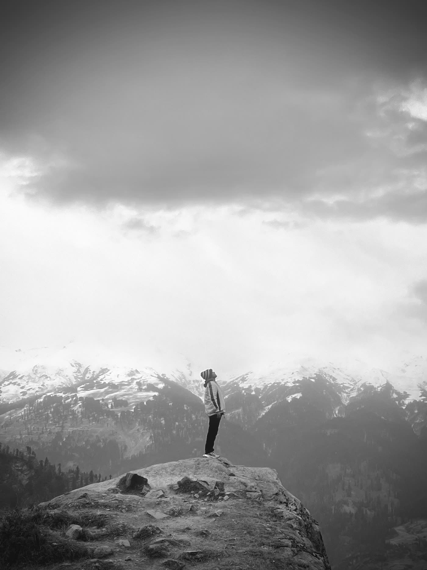 Tirth Raval standing on a rock ledge above a forested valley, snow peaks beyond